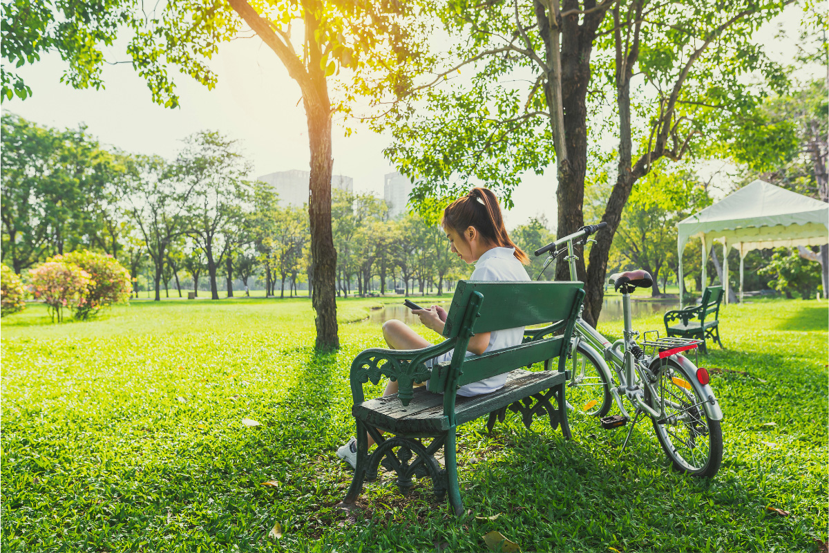 woman in sports attire sitting on a bench in an urban park with bicycle nearby checking her phone