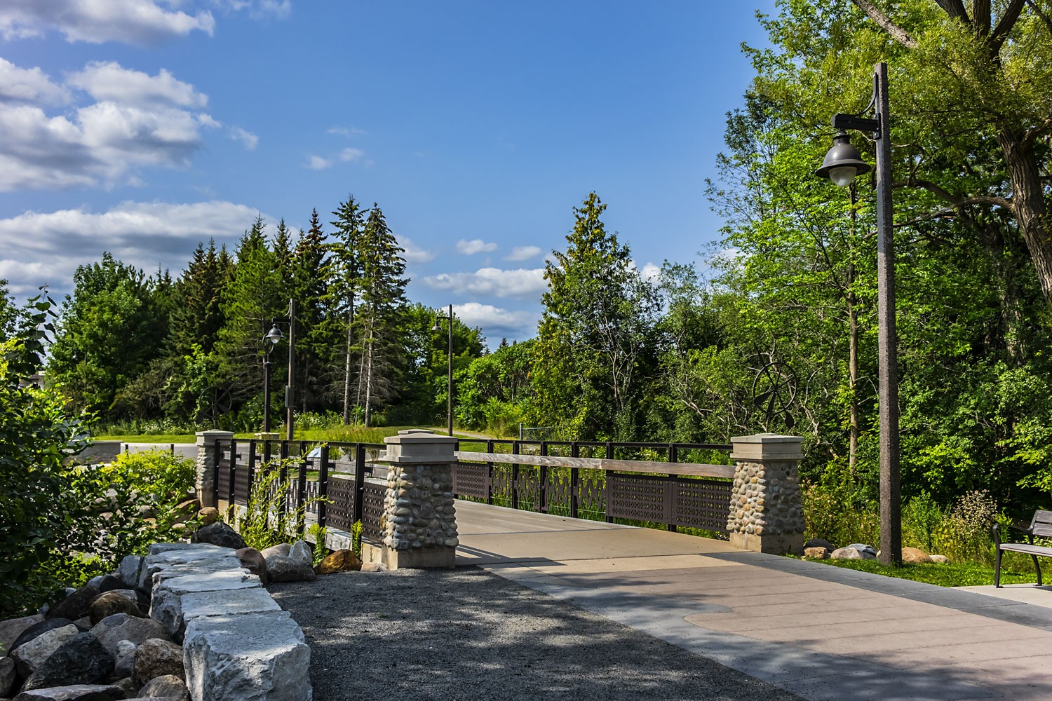 Paved pathway with a bridge, surrounded by trees on all sides with blue sky and clouds.