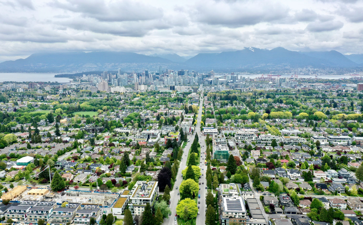 Vancouver from above showing city streets lined with trees, parks, the ocean, and mountains with a cloudy sky above.