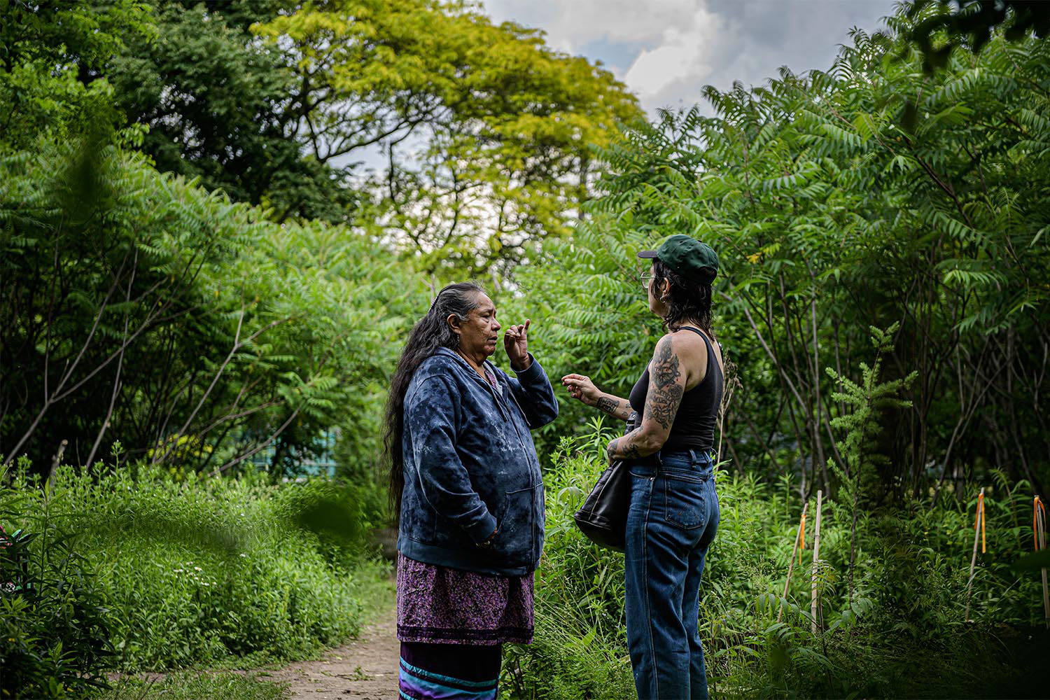 Indigenous woman and a tattooed woman with glasses stand in a forest together. Cloudy sky in the background.